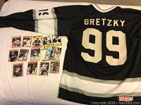 Photo of Wayne Gretzky black CCM Los Angeles Kings jersey back side with name 'GRETZKY' and number 99, and 14 hockey collector cards laid out beside the jersey.