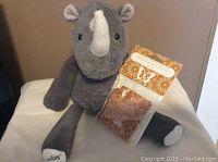 Front view of gray rhino plush sitting with white horn, black eyes, and fabric textures beside sealed scent pak labeled Rum Raisin Cookie with floral design.