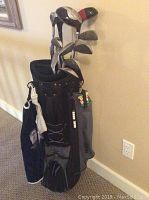 Photo of black golf bag holding set of women's right handed golf clubs with attached accessories (towel, tee holder) against beige wall.