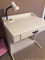 White wooden desk or vanity with a drawer featuring a decorative brass-colored handle, an open shelf beneath for storage, accompanied by a white office desk lamp with a flexible neck and round base placed on the top surface.
