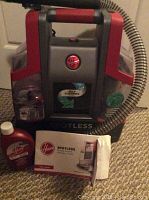 Front view of the Hoover Spotless portable carpet and upholstery cleaner, hose attached, cleaning solution bottle, and user manual in front.