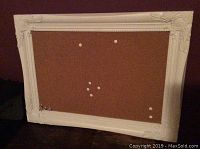 White framed hanging cork board with decorative ornamental corners, showing cork surface with some pin marks.