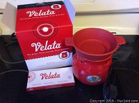 Image showing the red ceramic Velata fondue maker placed on a stovetop with its original branded box and a box of fondue forks beside it.