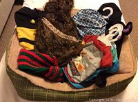 Small dog bed filled with various small dog clothes and costumes including the Chewbacca, Bumble Bee, and Dragon costumes, a rain jacket, t-shirts, and a checkered shirt.