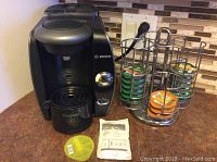 Black Bosch Tassimo coffee maker with metal carousel stocked with green and orange coffee pods next to it. Yellow descaling disk and descaling powder sachet placed in front.