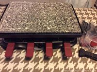 Top view of raclette grill with granite cooking surface and eight raclette dishes underneath with red handles, along with spatulas and accessories in plastic bags.