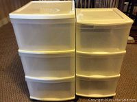 Photo showing front and side view of two white plastic rolling storage carts with three drawers each, placed side by side on carpeted floor.