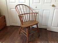 Side angle view of the wooden Windsor chair showing curved back, vertical spindles, worn seat, repaired right arm and leg, set in a room with wood floor and white doors.