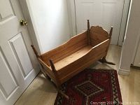 Side/front angled view of the antique pine crib on a red patterned rug, showing the rocking curved base and turned posts with finials.