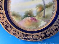 Full view of the hand painted plate showing the central idyllic scene and heavy gilded border with a deep blue background.