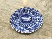 Full view of the round blue and white ceramic charger plate with intricate pattern and Greek key border, placed on a marble surface.