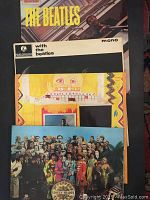 Four vintage Beatles vinyl records stacked, showing colorful front covers including Sgt. Pepper's and With The Beatles mono album.