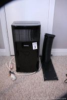 Front view of black portable air conditioner with attached power cord, alongside flexible black exhaust hose and vent panel.