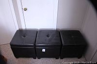 Three dark brown faux leather cube storage ottomans lined up side by side on carpeted floor in front of white door.