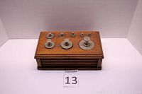 Oak box with metal weights displayed on top, showing nine weights of varying sizes arranged from smallest to largest.