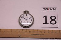 Front view of Illinois pocket watch showing white dial with black Arabic numerals, blue hands, and subdial at 6 o'clock next to ruler for scale.