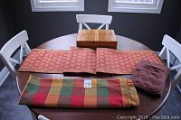 Four-piece lot on table including art deco wooden box, red patterned table runner, plaid table cloth folded, and brown fabric remote control holder.