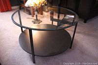 Front angled view of the round coffee table showing the glass top, metal legs, and wooden lower shelf on carpet.