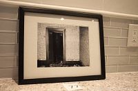Framed black and white photograph showing a doorway in an abandoned room with peeling wall texture and debris on the floor.
