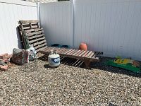 Wide view of garden bench, pallets, propane tank, soil and fertilizer bags placed on gravel in outdoor fenced area.