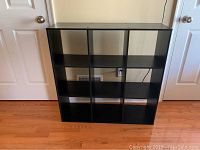 Front view of black cubby storage shelf with 9 square compartments on hardwood floor next to double doors.
