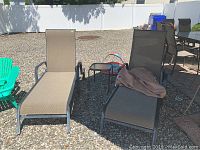 Two outdoor wheeled lounge chairs on gravel with one small glass topped side table between them and one brown chair cover draped over one chair.