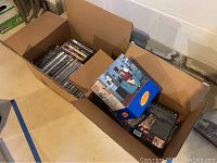 Two cardboard boxes with DVDs, one smaller box contains a Seinfeld box set with no DVDs, script copy and accessories inside, other box contains various DVDs including some sealed.