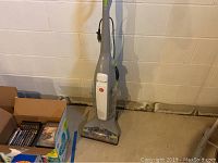 Full view of the Hoover Floormate Deluxe floor cleaner showing tall handle and base unit on a basement floor near a box of household items.