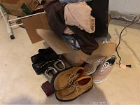 Overview of assorted men's shoes, boots, and clothing including folded fabric items, shoes of black, tan, and beige colors, some boxes in background