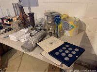 Table view showing silverplated urn, table linens, medals, lampshade, and other items on white folding table in basement
