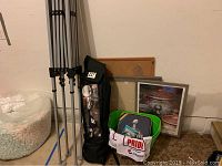 Photo showing folded EZ Up canopy with black carrying bag featuring NY Giants logo, green tub, framed pictures, and message board leaning against wall.