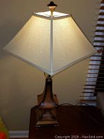 Full view of the table lamp showing wooden sculpted base, pyramid cream fabric shade, and brass finial.
