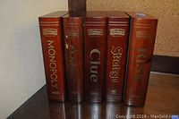Five wooden boxes standing upright, spine view showing game titles Monopoly, Scrabble, Clue, Stratego, and Risk in a vintage-style design.