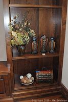 Photo showing three metal vases with faux flowers and three decorative candle holders displayed on a wooden shelf, with a decorative bowl containing natural sphere-like objects below.