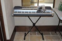 Full view of Casio LK90 keyboard on black X-style stand against wall under window.