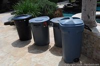 Four outdoor trash cans arranged on patio tile flooring near a pool and plants.