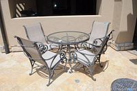 Patio set with round glass top table and four metal framed mesh chairs arranged outdoors on stone patio.