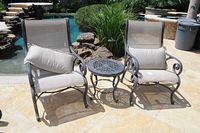 Two metal patio chairs with mesh seats and backs flanking a round metal coffee table, each chair includes a beige pillow. Outdoor setting with stone patio and greenery.