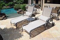 Two patio lounge chairs with light taupe mesh fabric and dark metal frames, one chair has a large rip in the fabric seating. Small round metal coffee table between the chairs.