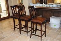 Three wooden bar stools with leather seats arranged in a kitchen setting, showing overall condition and style.