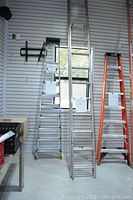 Photo showing three ladders against a wall: an aluminum step ladder, a 21 ft aluminum extension ladder, and an orange fiberglass step ladder.