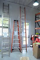 Three ladders standing upright against a wall: two silver metal extension ladders (one 8ft, one 10ft) and one orange metal step ladder approximately 6ft tall.