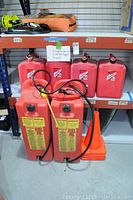 Shelved four 5-gallon red gas cans stacked behind two tall 11-gallon red Fuel Pal cans with black hoses and four orange cones beside them.
