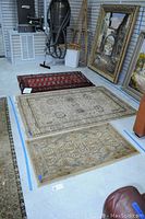 Three rugs laid out on floor, size and color differences visible, surrounded by framed paintings and some equipment
