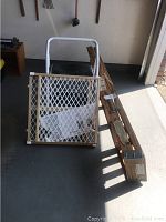 Photo showing a wooden mesh child safety gate standing upright and a long wooden step ladder lying on the ground.
