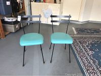 Pair of retro kitchen chairs showing front view with turquoise vinyl seats and black metal frames with gold feet caps.