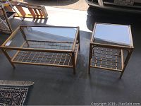 Pair of rattan coffee tables with rectangular clear glass tops and lattice rattan lower shelves shown side by side on concrete floor with rugs and nearby furniture.