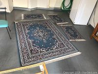 Four blue wool carpets arranged on a concrete floor including one large and three smaller carpets, all with floral medallion patterns and fringed edges.