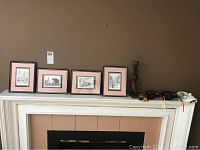 Four framed New Orleans prints with pink mats lined on mantel, plus two ceramic masks and wood figurine to the right.