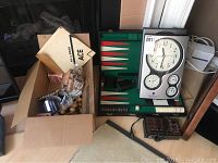 Box of assorted small items including wood spheres, a measuring tape, and other small items alpha, next to backgammon set and battery clocks.